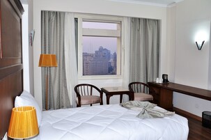 Habitación de hotel con cama, escritorio, dos sillas, lámpara y ventana con vista a la ciudad.