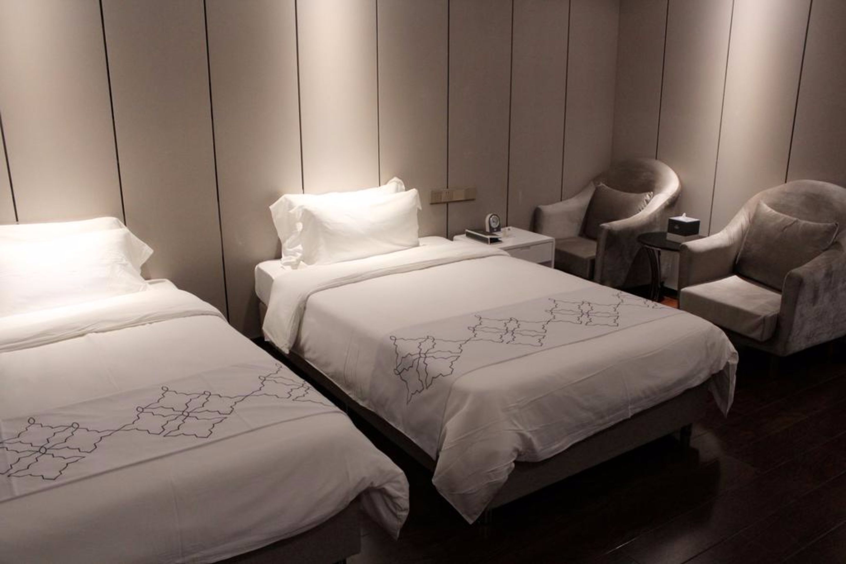 A hotel room with two beds, a sitting area with a chair, a bedside table, and a small table with a lamp.