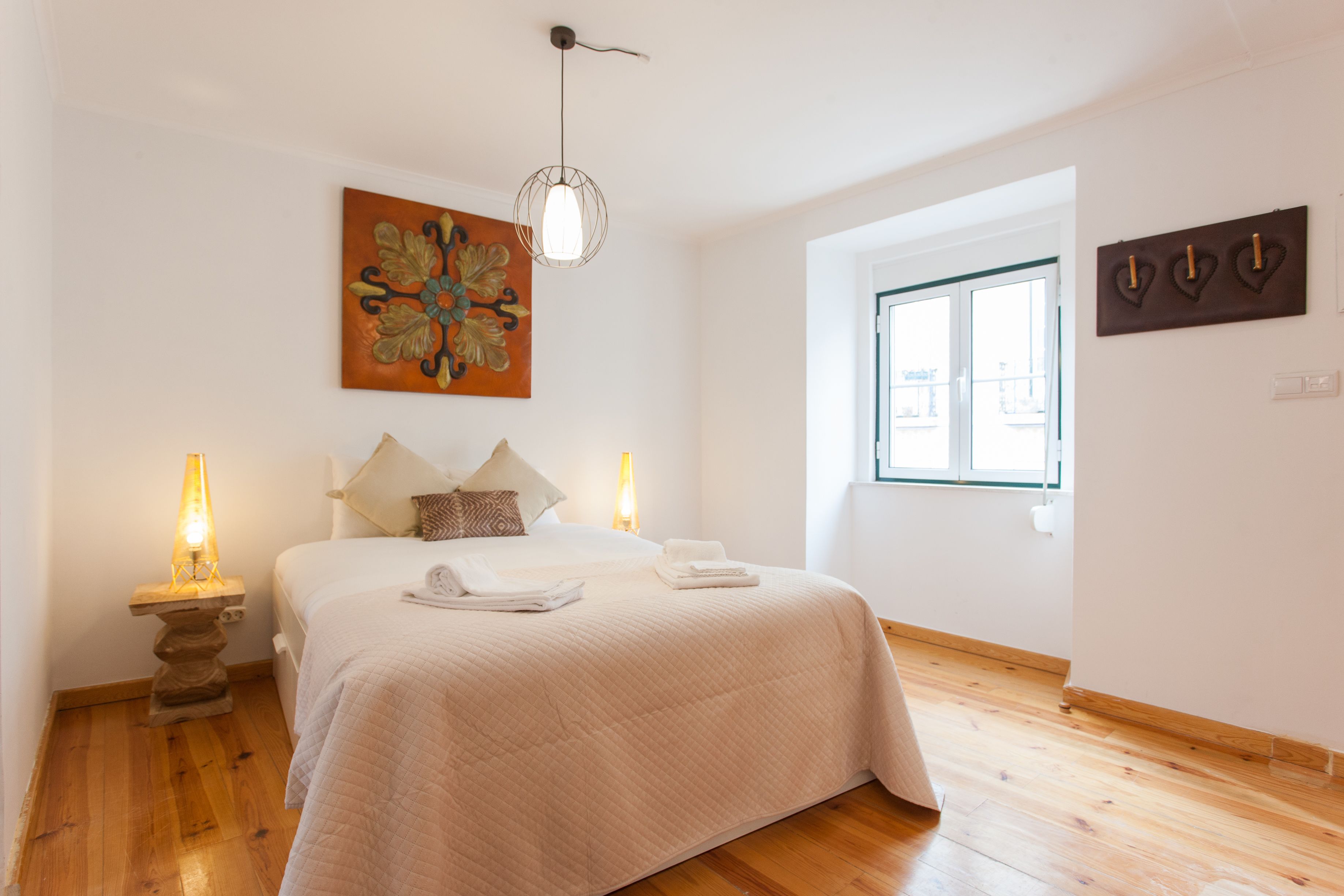 Un dormitorio con una cama, dos lámparas de noche, un cuadro, una ventana y una lámpara fijada a la pared.