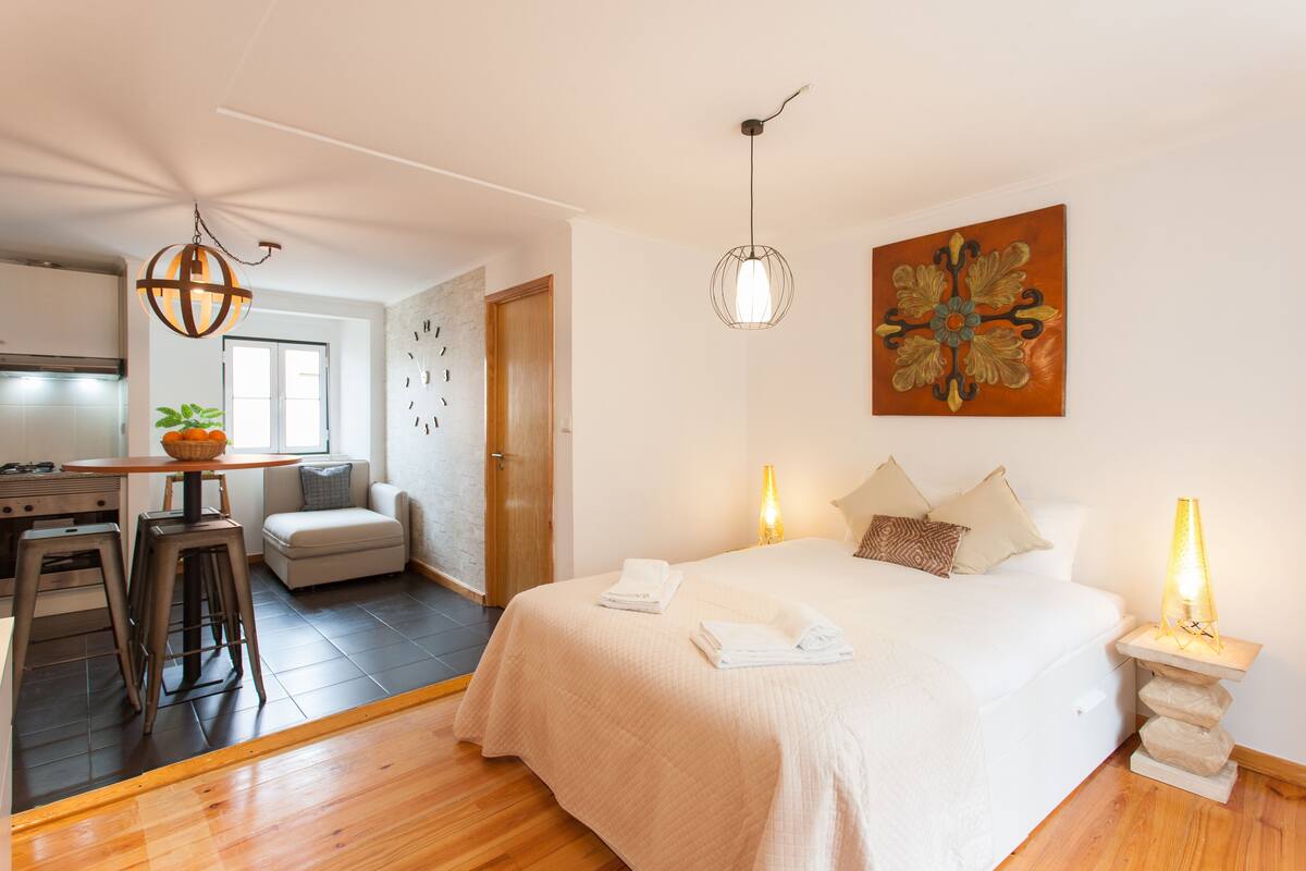 A modern bedroom with a large bed, bedside lamps, and a dining area with stools.