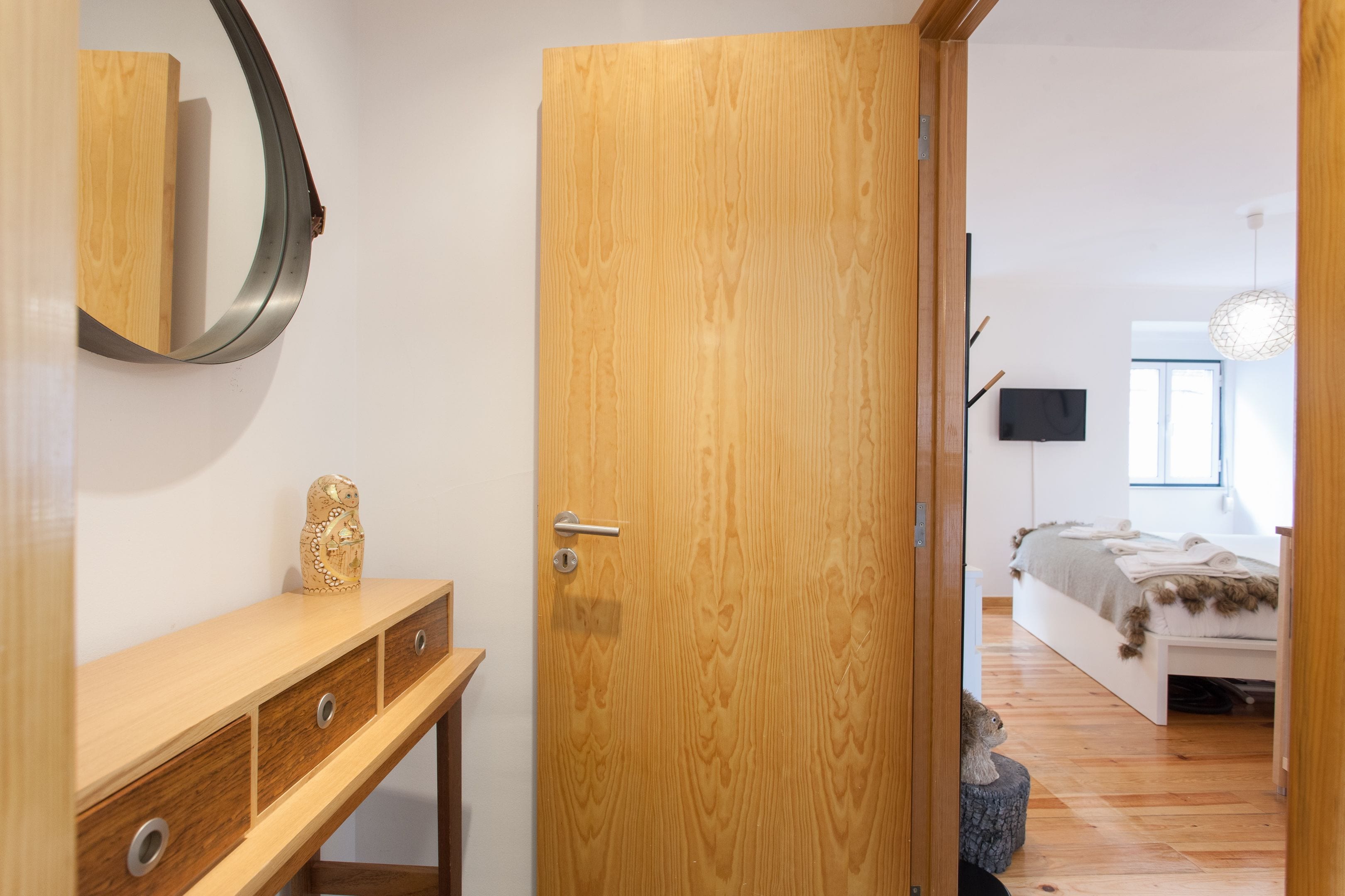 Una porta de fusta que dona a un dormitori amb un llit, una taula de nit i un televisor penjat a la paret.