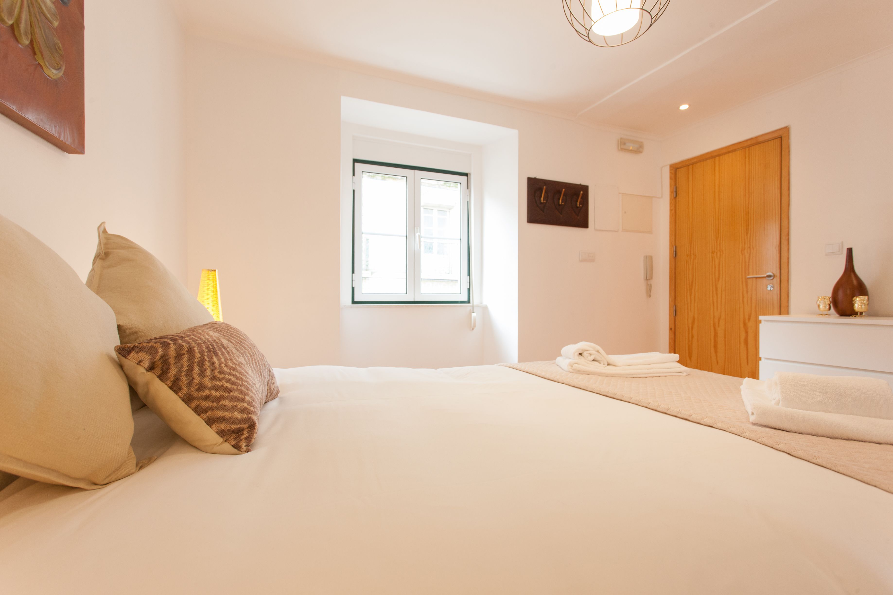 Une chambre à coucher avec un lit, des oreillers, une fenêtre, une porte en bois et une table de chevet sur laquelle se trouve un vase.