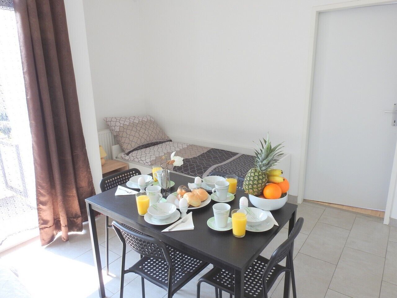 A dining table set with plates, bowls, and fruits, with a bed and curtains in the background.