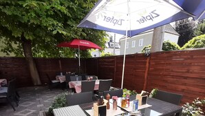 An outdoor patio with a glass-top table, wicker chairs, and red and blue umbrellas. - Apartments CITY Stadion (Linz)