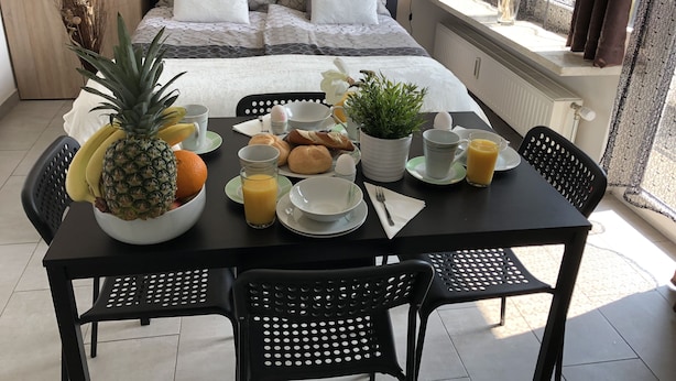 A breakfast setup with a bed in the background, a table with breakfast items, and a window with curtains. - Apartments CITY Stadion (Linz)
