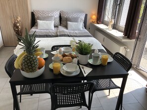 A breakfast setup with a bed in the background, a table with breakfast items, and a window with curtains. - Apartments CITY Stadion (Linz)
