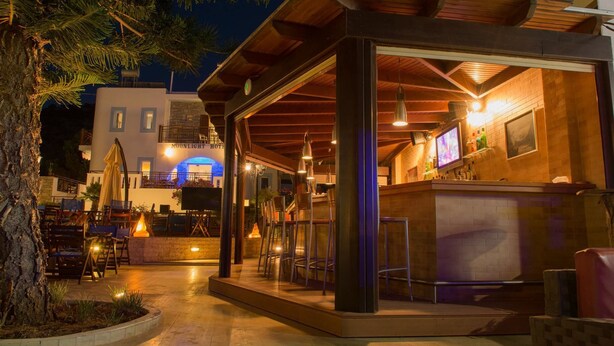 A cozy outdoor bar area with wooden seating and a bar counter, surrounded by a stone wall and a tree. - Moonlight Hotel (Hersonissos)