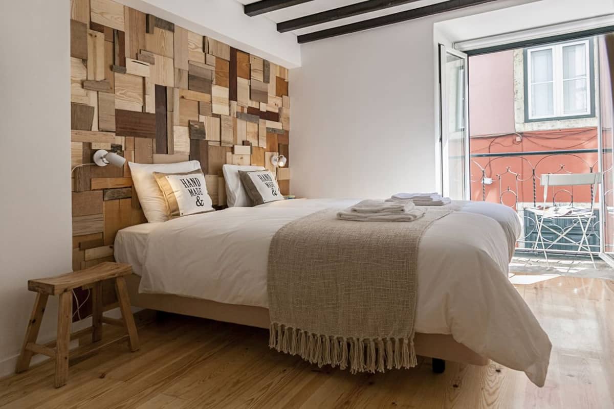 A bedroom with a wooden headboard, a bed with white linens, a small wooden stool, and a view of an outdoor balcony.