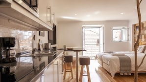 A modern kitchen with a black countertop, wooden stools, and a dining table. There is a bed with a wooden headboard and a window with a view of a building. - Bairro Studio Apartment - by LU Holidays (Lisbon)