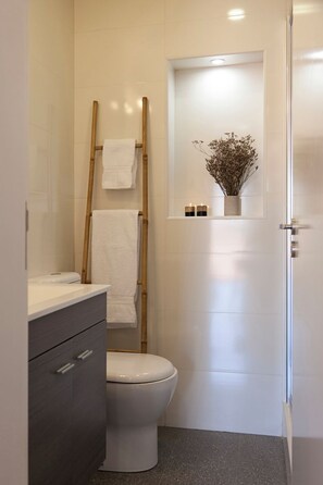 A bathroom with a toilet, a vanity with drawers, a towel rack, a shelf with a vase and candles, and a door with a towel hanging on it. - Bairro Studio Apartment - by LU Holidays (Lisbon)