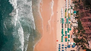 An aerial view of a beach with rows of umbrellas and beach chairs, a rocky cliff on one side, and waves crashing onto the shore.