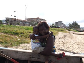 Una mujer sostiene a un niño en una playa arenosa, con edificios al fondo.