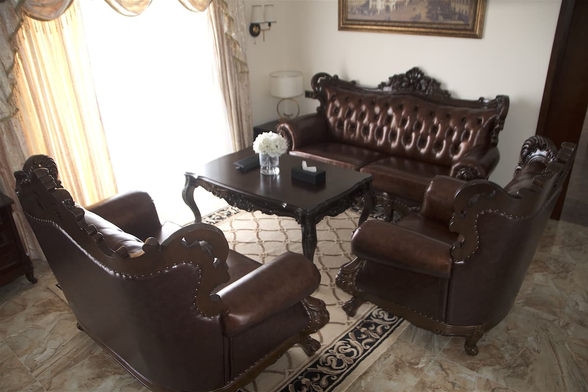 Ein Zimmer mit einem braunen Ledersofa, zwei passenden Sesseln, einem kleinen Tisch mit einer Vase und einer Taschentuchbox, einer Lampe und einem gemusterten Teppich.