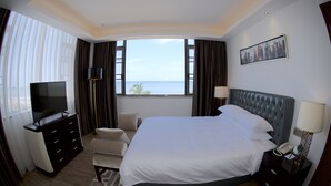 A hotel room with a large bed, a TV, a desk with a lamp, and a view of the sea.