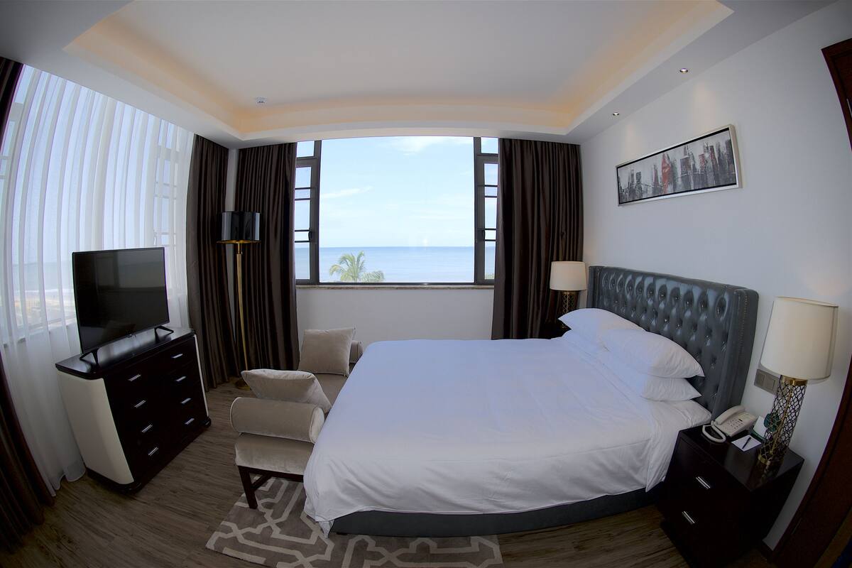 Ein Hotelzimmer mit einem großen Bett, einem Fernseher, einem Schreibtisch mit Lampe und Meerblick.