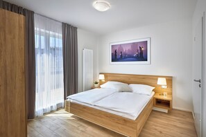 Une chambre d’hôtel moderne avec un lit en bois, une table de chevet avec une lampe, un tableau encadré au mur et une grande fenêtre avec des rideaux.