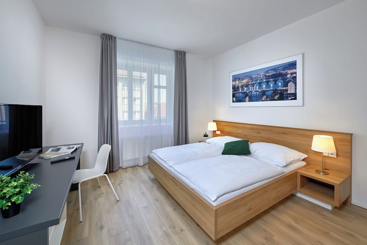 Een hotelkamer met een bed, nachtkastje, lamp, stoel, bureau, televisie en een raam met gordijnen.