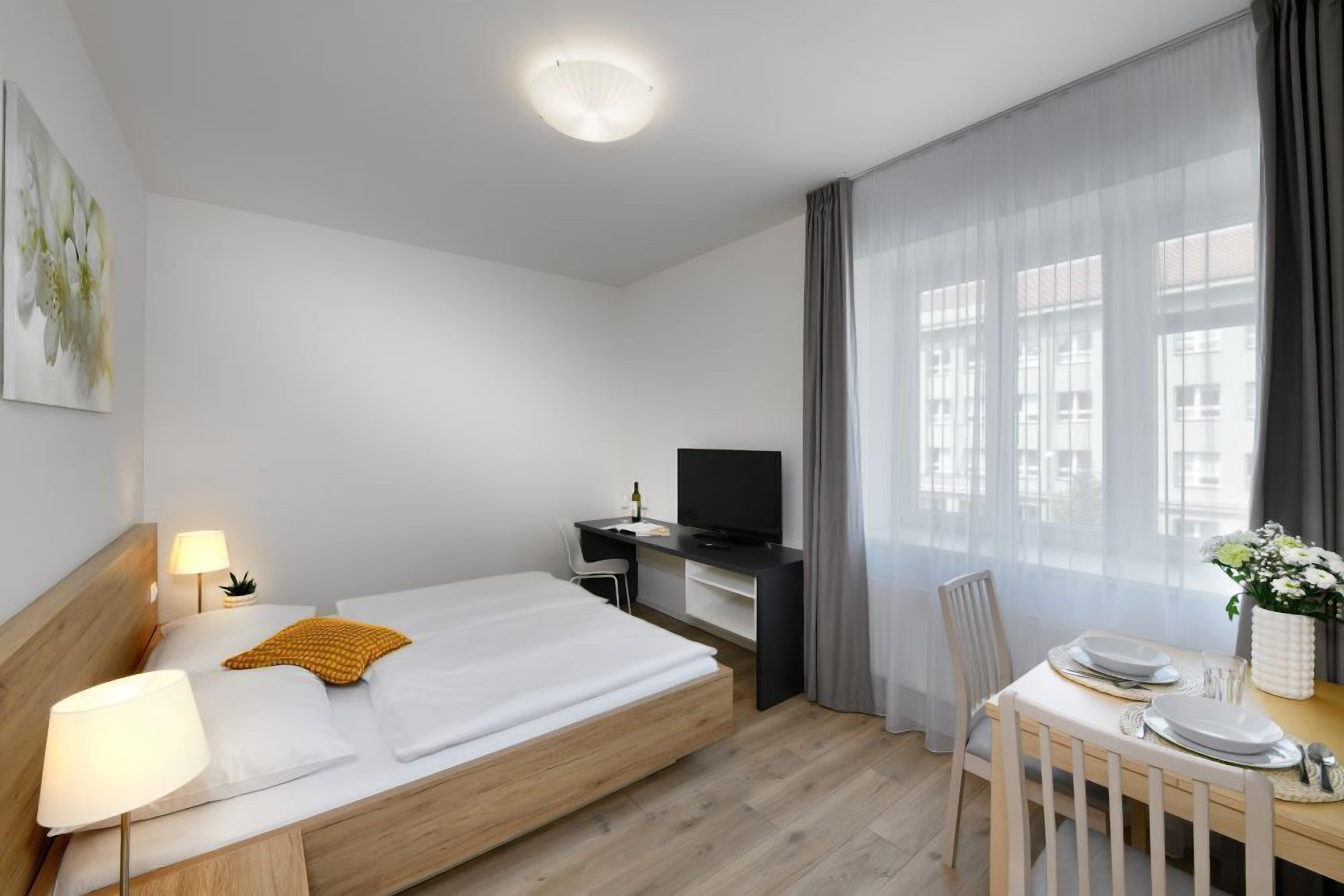 Una habitación de hotel moderna con una cama de madera, un escritorio con televisor, una mesa de comedor y un gran ventanal con cortinas translúcidas.