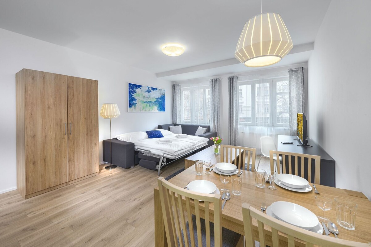 A modern hotel room with a bed, a dining table set for two, a wardrobe, and a large window with curtains.