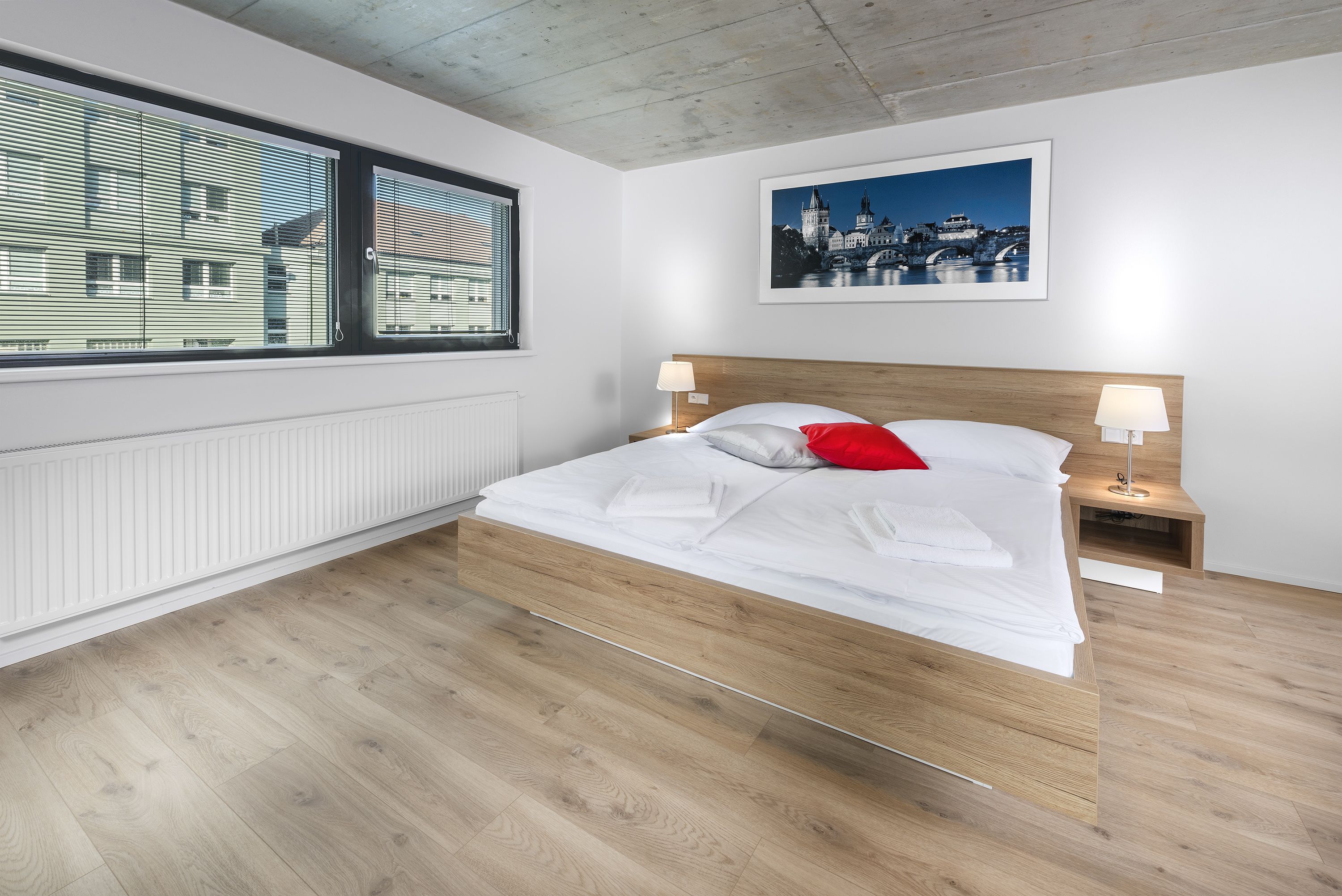 A modern bedroom with a wooden bed, white bedding, a red pillow, and a cityscape view through the window.
