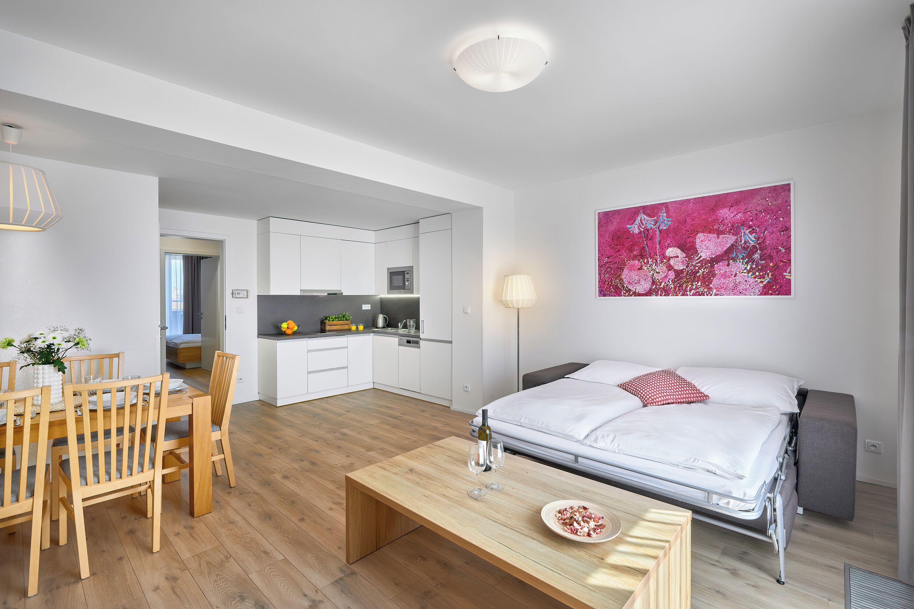 Una stanza moderna e luminosa con un tavolo da pranzo in legno, un letto con biancheria bianca, una zona cucina e un'opera d'arte rosa vivace appesa al muro.