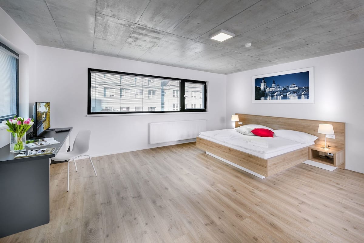 Een moderne slaapkamer met een houten bed, een bureau met een computer, een stoel, een raam met jaloezieën en een stadslandschap schilderij.