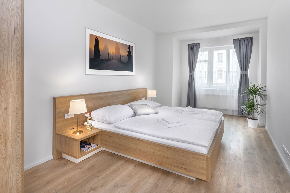 A modern hotel room with a wooden bed, bedside lamps, a framed picture, a window with curtains, and a potted plant.