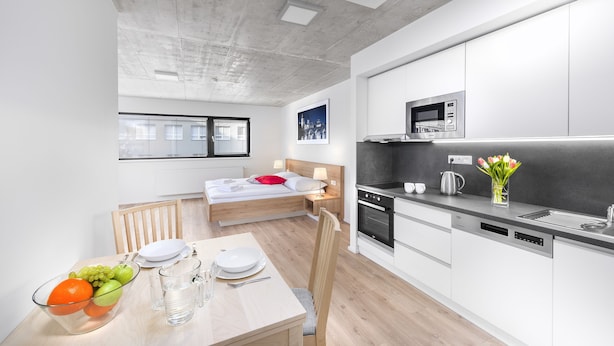 A modern kitchen with white cabinets, a microwave, and a dishwasher. There is a dining table with plates, glasses, and a fruit bowl. A bed with a red pillow is visible in the background.