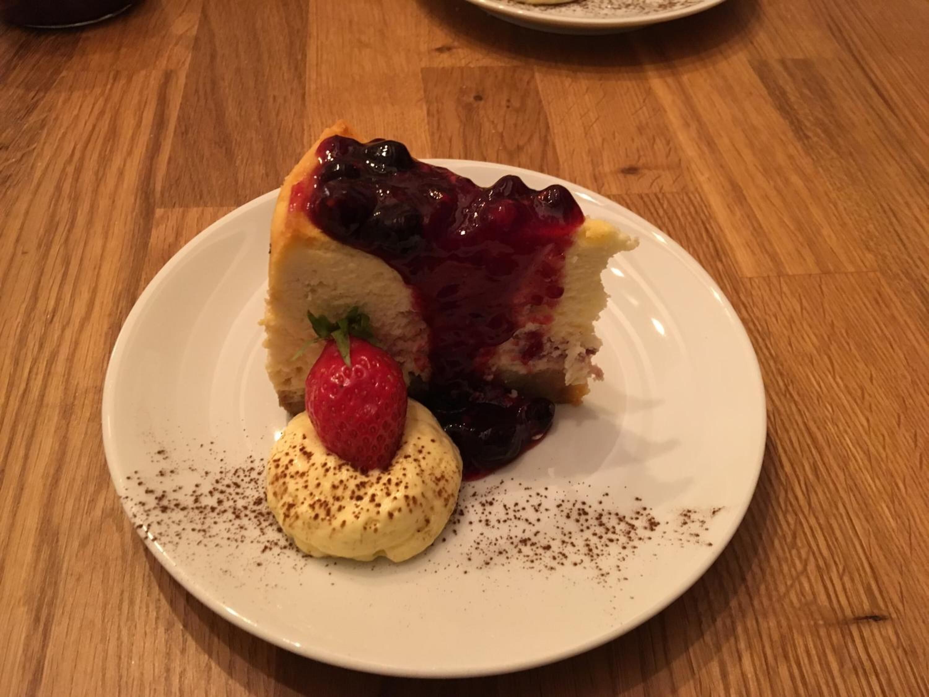 Een stuk cheesecake met bessensaus op een wit bord, vergezeld van een bolletje vanille-ijs en een aardbei.