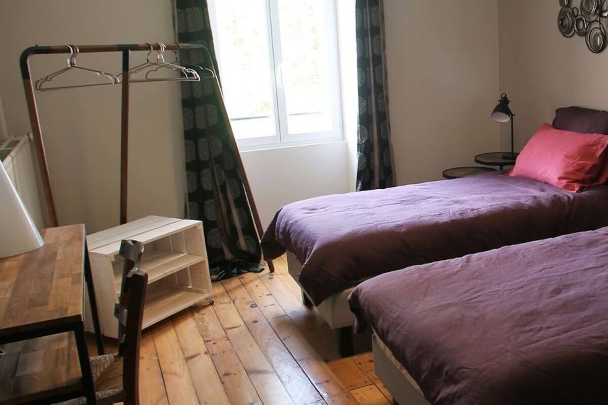 Une chambre avec deux lits, un plancher en bois, un porte-manteau, une table de chevet et une fenêtre avec des rideaux.