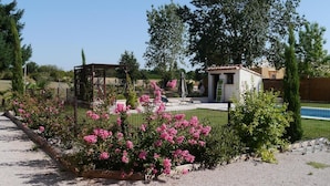 Un jardín con flores rosadas, un pérgola y un edificio pequeño con techo de tejas.