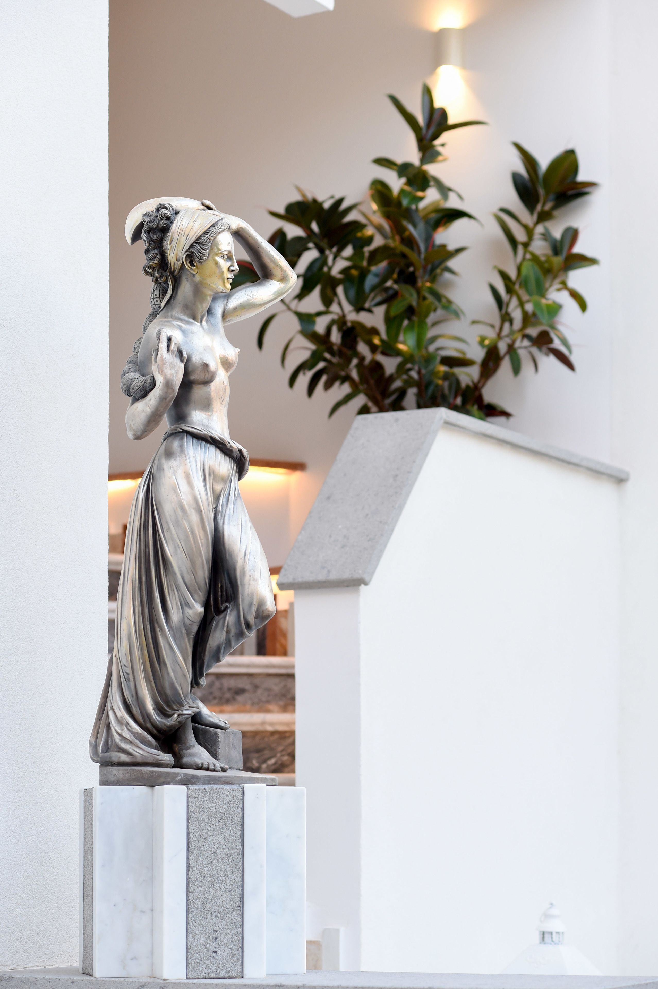 A statue of a woman in a classical pose, placed on a pedestal in a well-lit indoor space with a plant and a wall-mounted light fixture in the background.