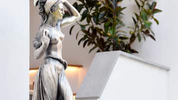 Une statue d’une femme dans une pose classique, placée sur un piédestal dans un espace intérieur bien éclairé, avec une plante et un luminaire fixé au mur en arrière-plan.