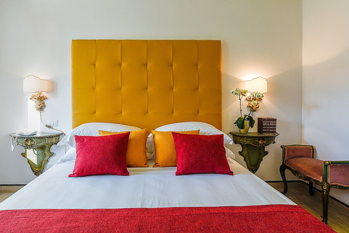 Una habitación de hotel con una cama grande, dos mesitas de noche con lámparas, una silla y una mesita con una planta.