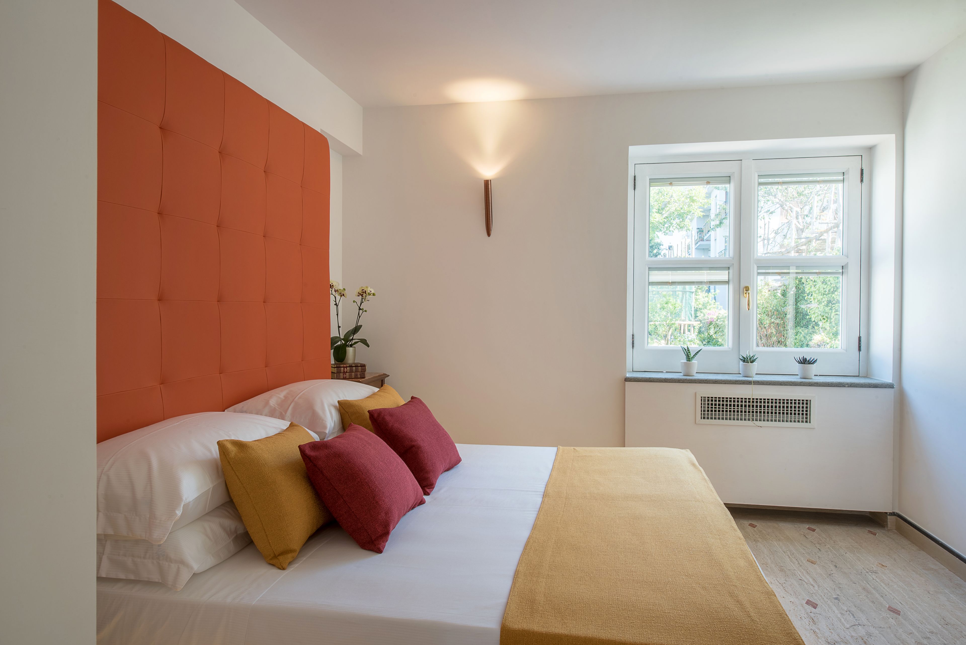 A bedroom with a bed, pillows, a bedside table with a plant, and a window with a view of trees.
