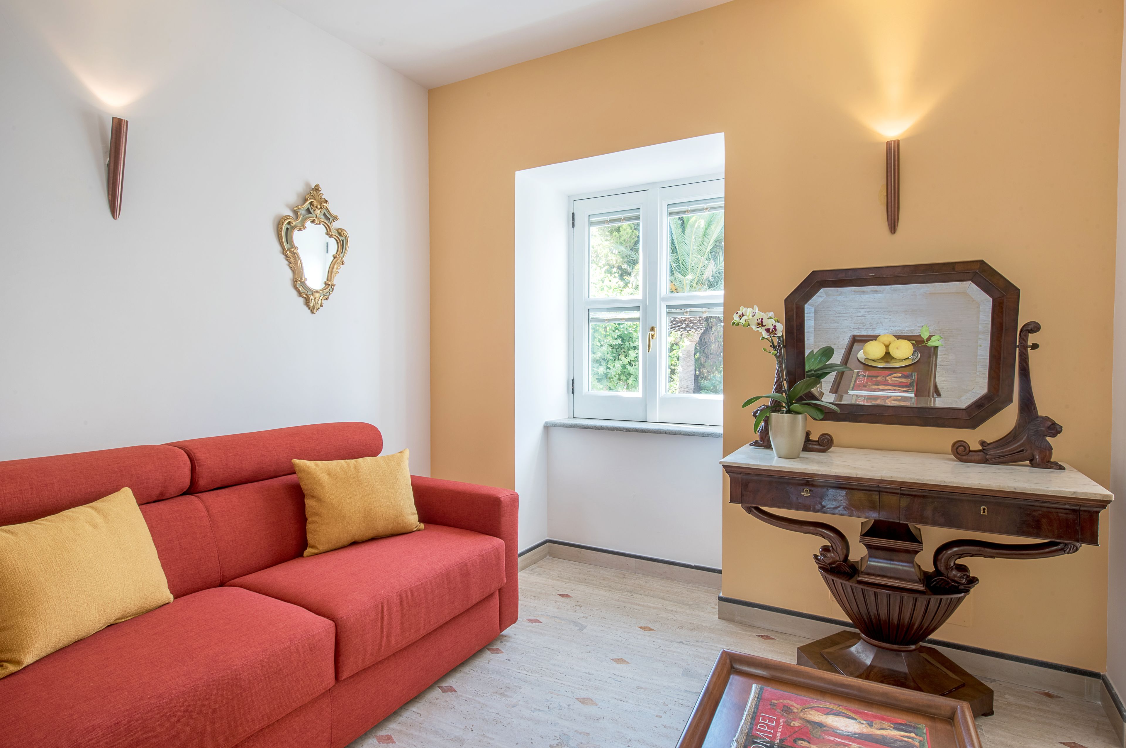 Un salon avec un canapé rouge, une petite table surmontée d’un miroir et une fenêtre donnant sur un paysage verdoyant.
