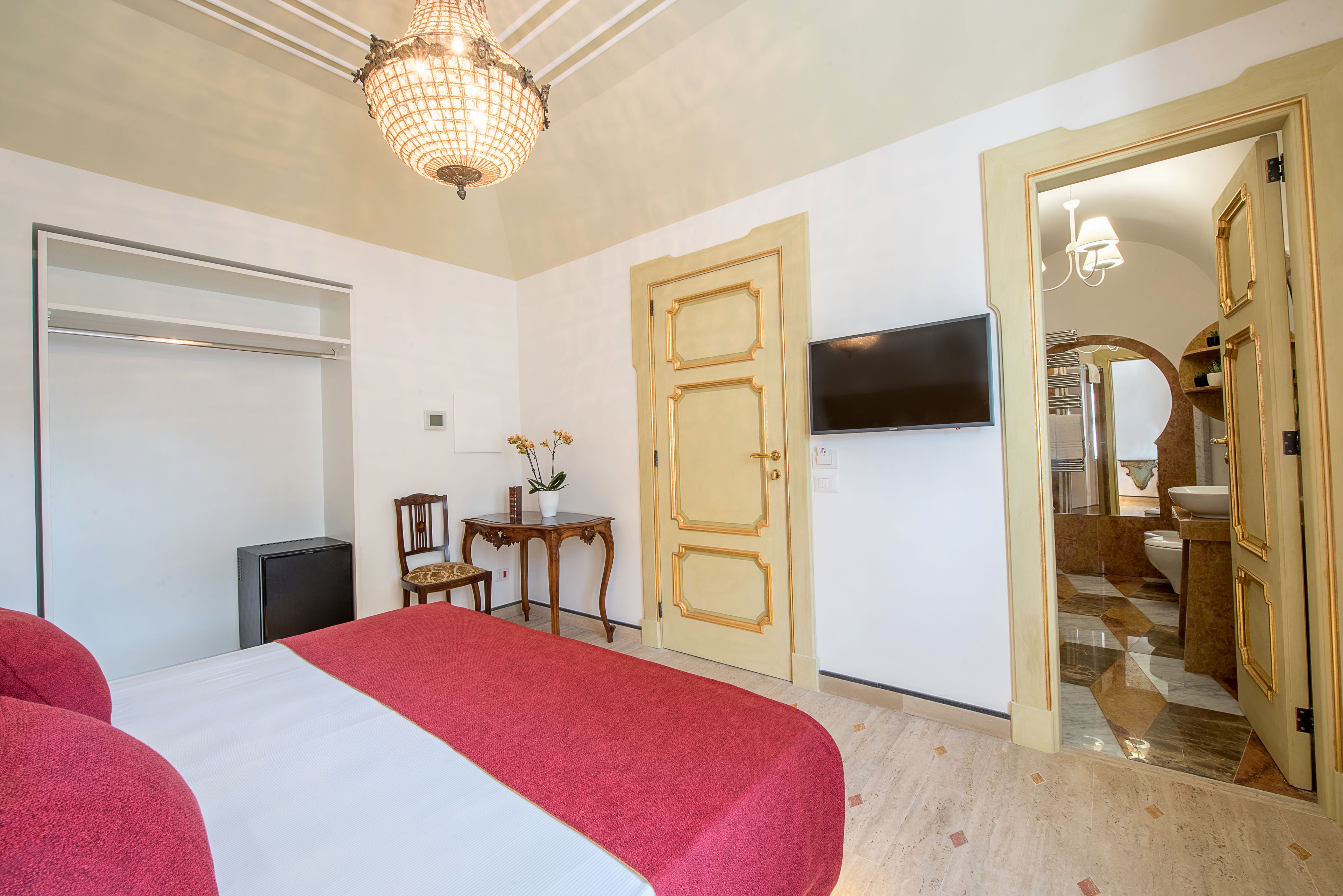 Habitación de hotel con cama, televisión, puerta, una mesita con una planta y un candelabro.