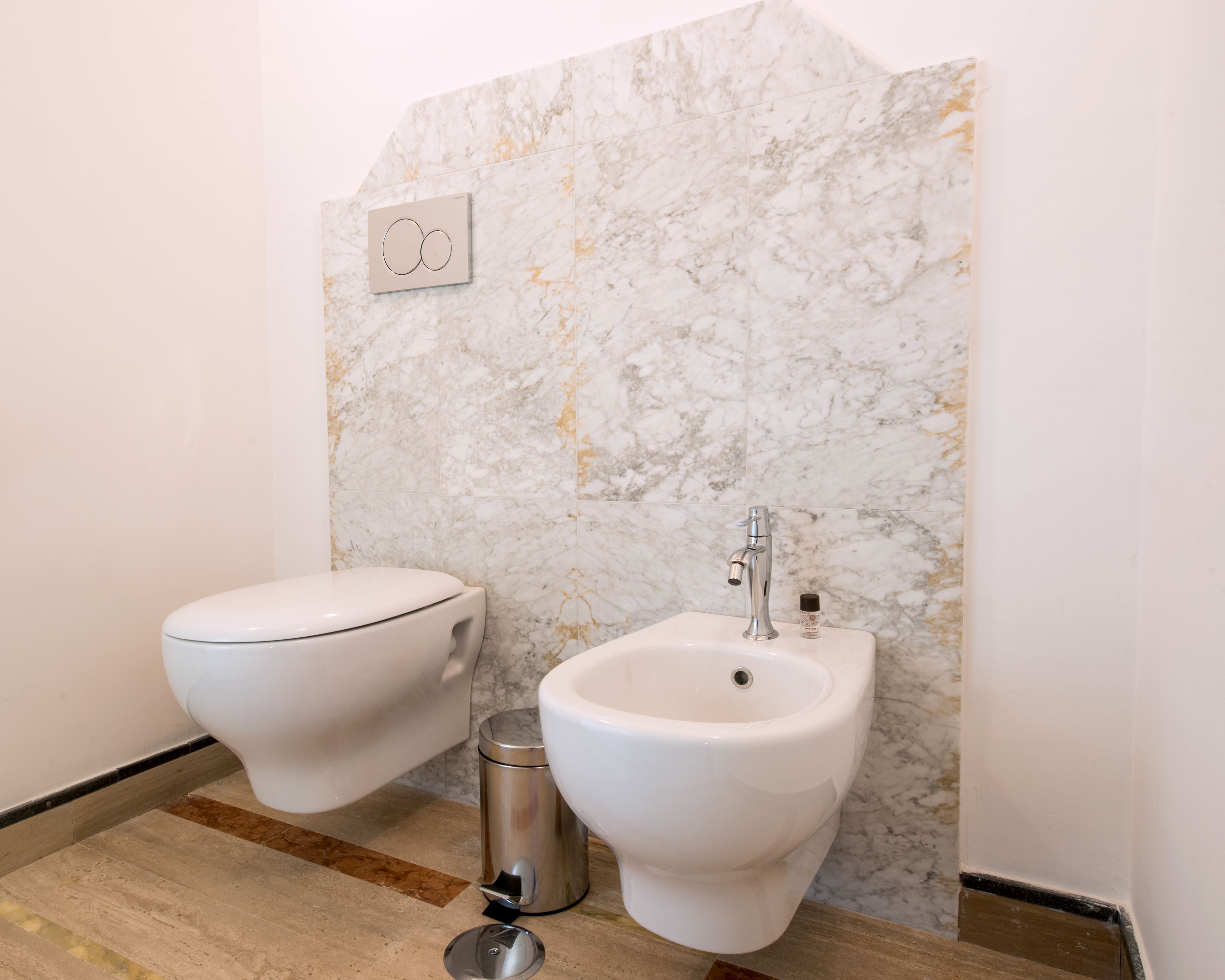 A bathroom with a marble wall, a toilet, a bidet, and a trash bin.