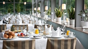 A long dining table set with breakfast items, including fruits, pastries, and beverages, in a well-lit room with large windows and potted plants. - Schloss Berge (Gelsenkirchen)