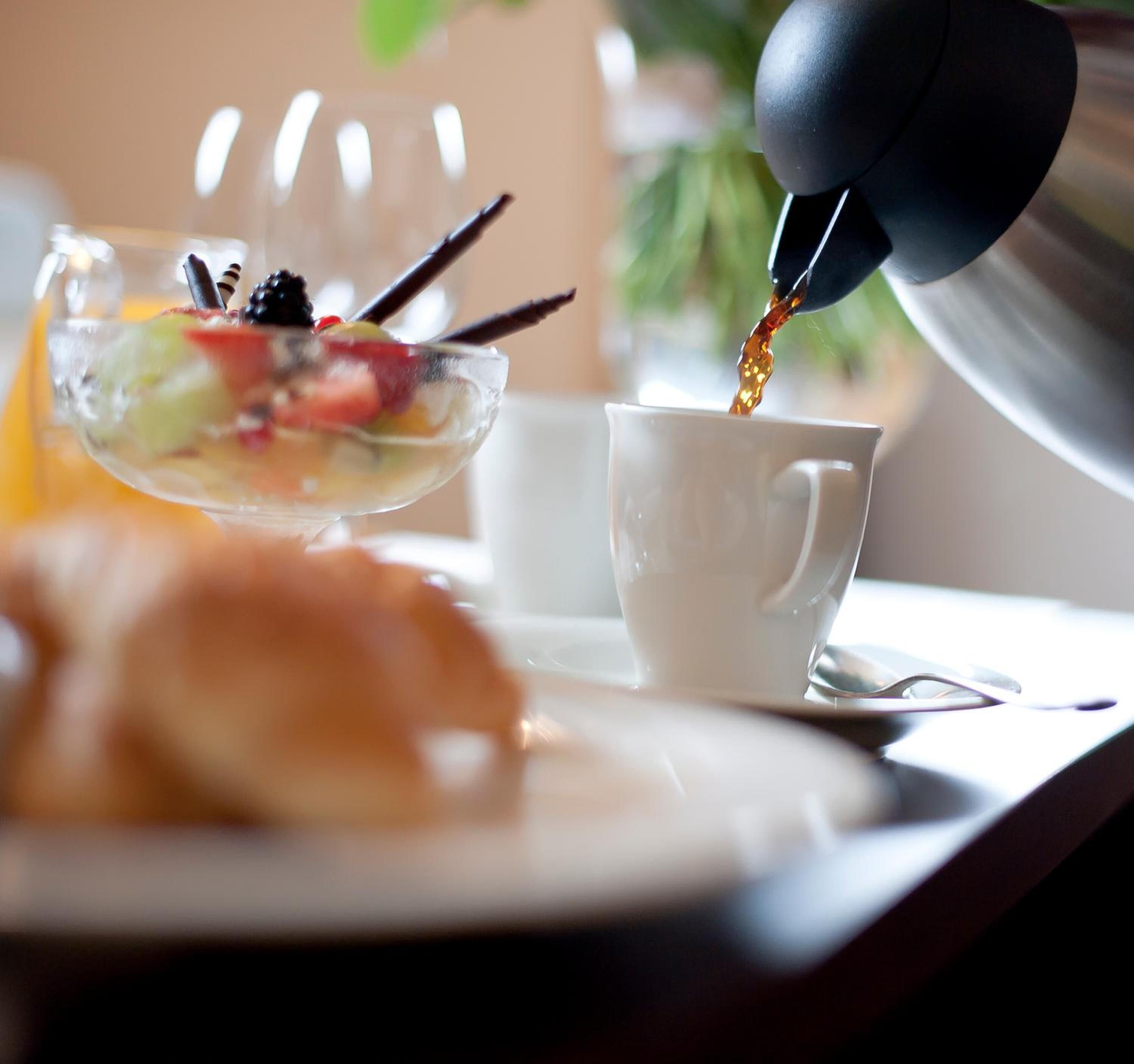 Una máquina de café vertiendo café en una taza blanca, un cuenco con fruta y un croissant sobre la mesa.