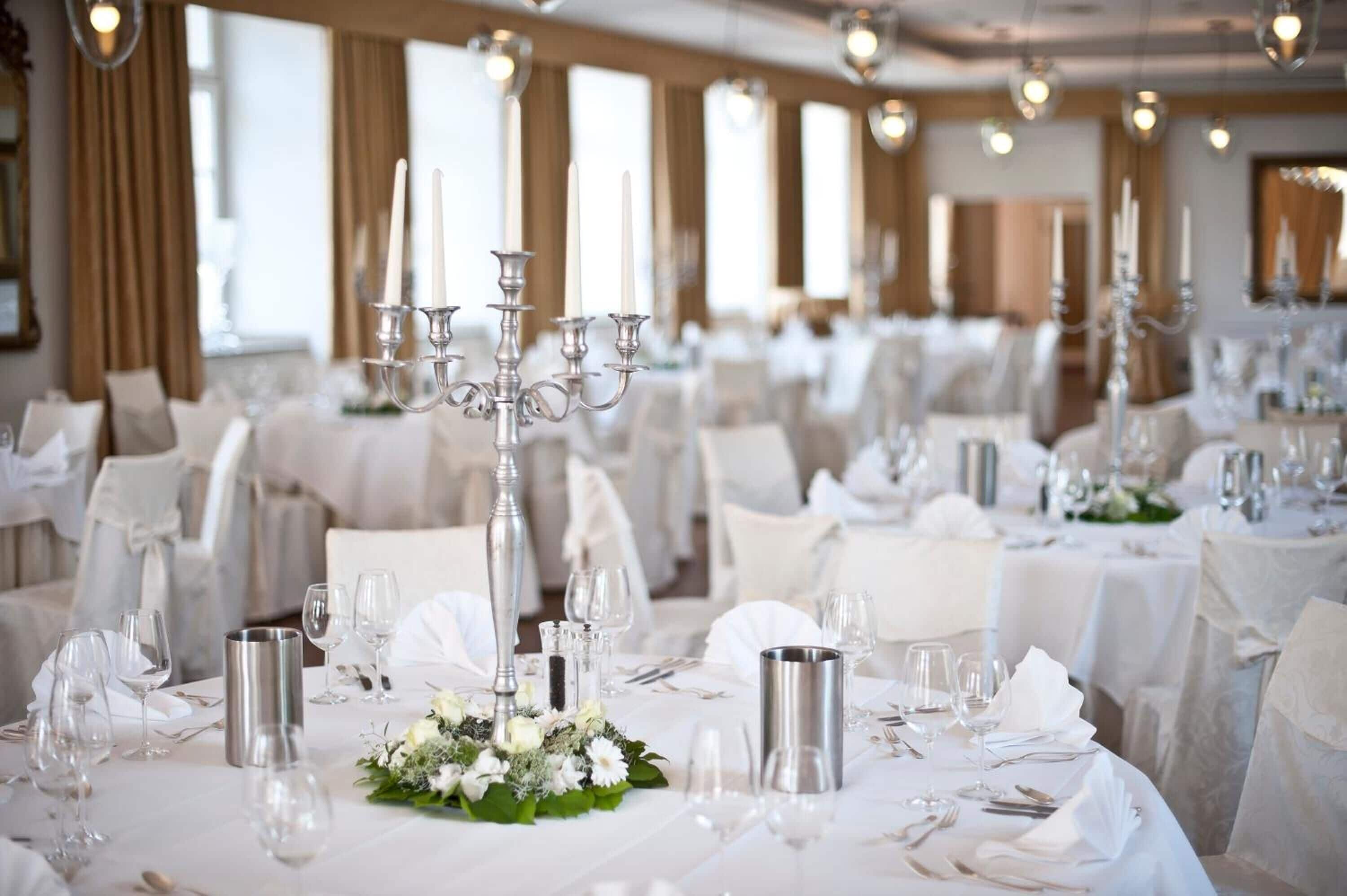 Ein Festsaal mit runden Tischen, festlich gedeckt für eine formelle Veranstaltung, mit Kerzenständern, weißen Tischdecken und Blumenschmuck in der Mitte.
