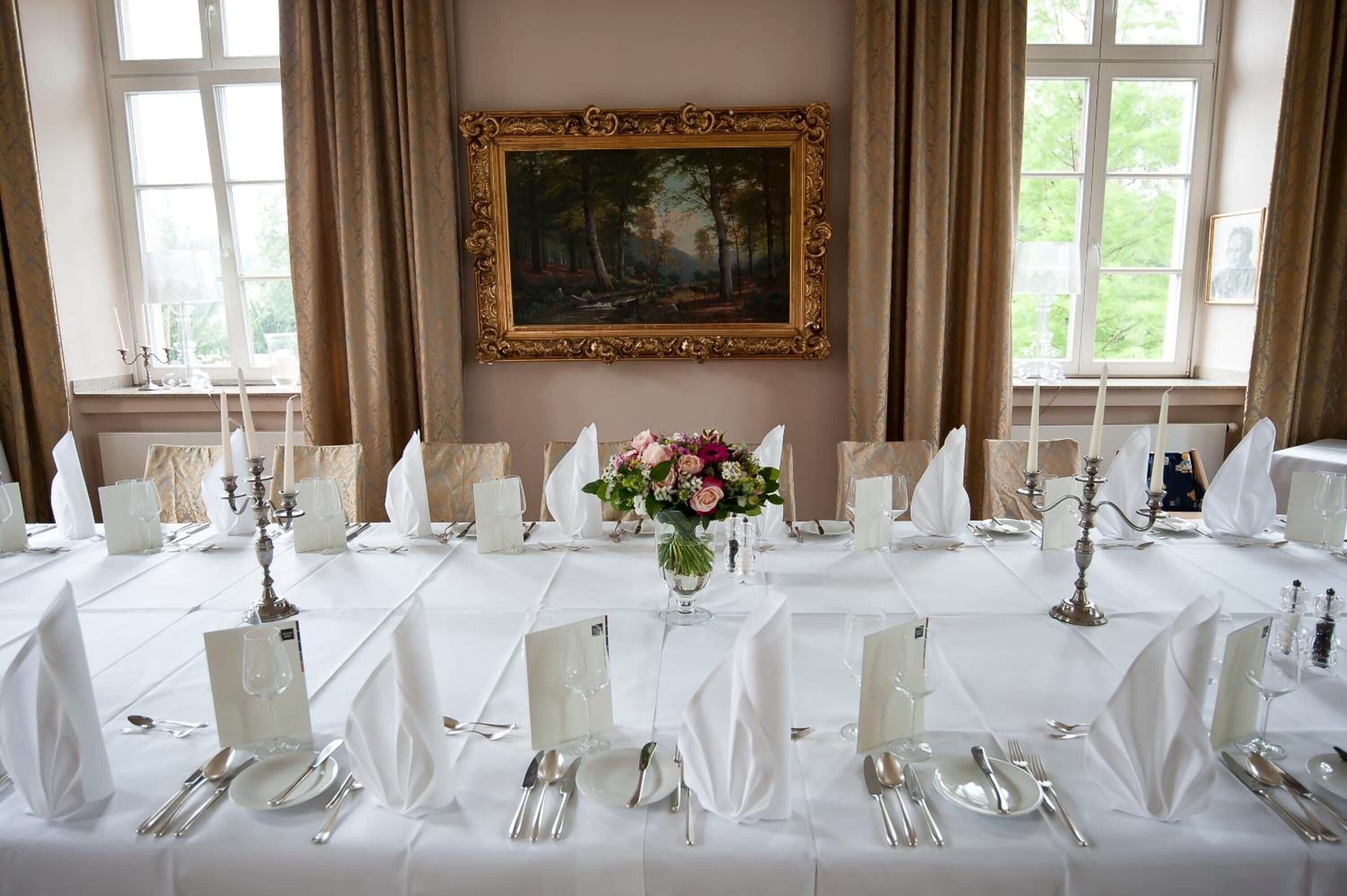 Et elegant spisebord dækket med hvide duge, sølvtøj og lysestager, med et maleri og en vase med blomster på væggen bagved.