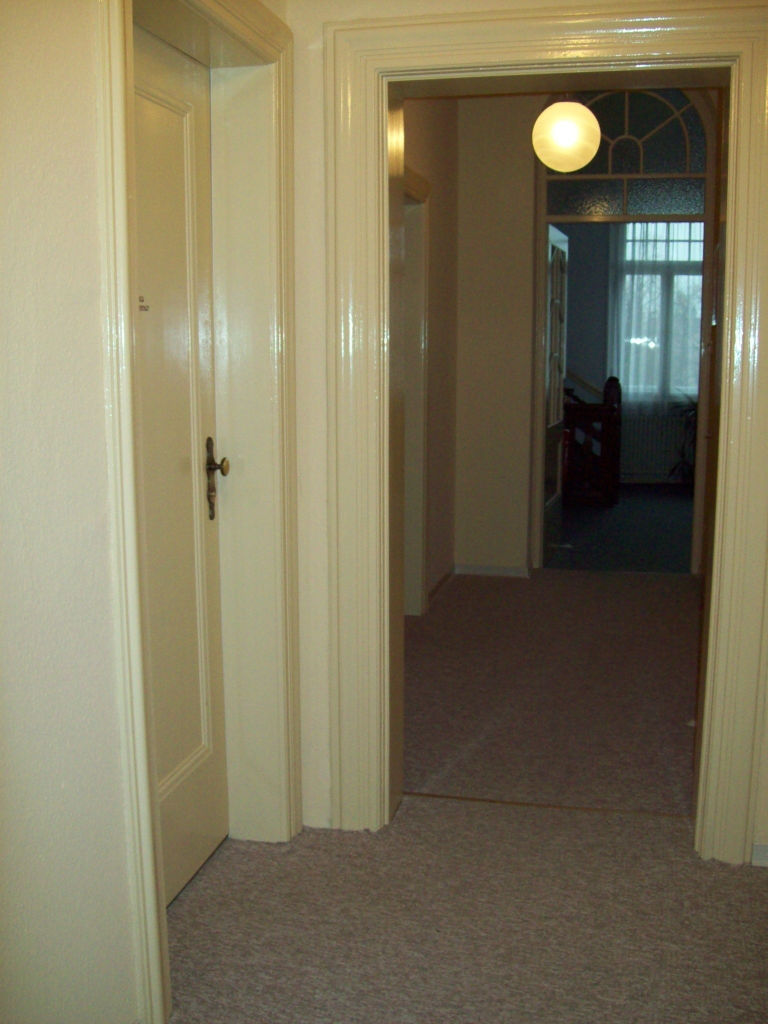 A hallway with a carpeted floor, a door on the left, and a window with a light fixture in the background.