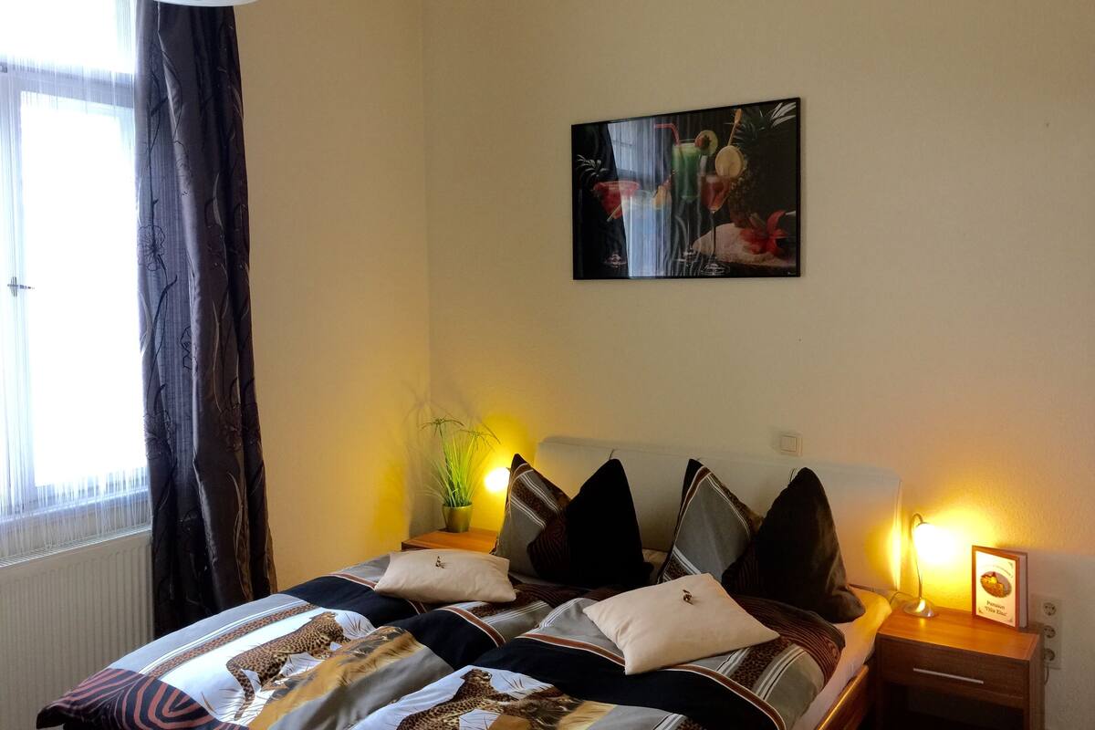 A hotel room with a bed, two bedside tables, a nightstand, a potted plant, a painting, and a window with curtains.