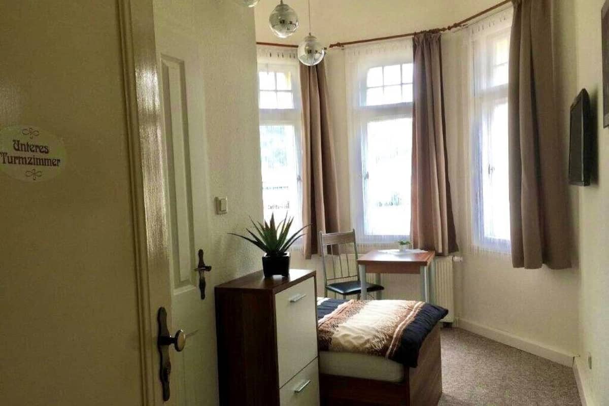 A room with a bed, a desk, and a chair. There is a window with curtains, a plant on the desk, and a sign on the door.