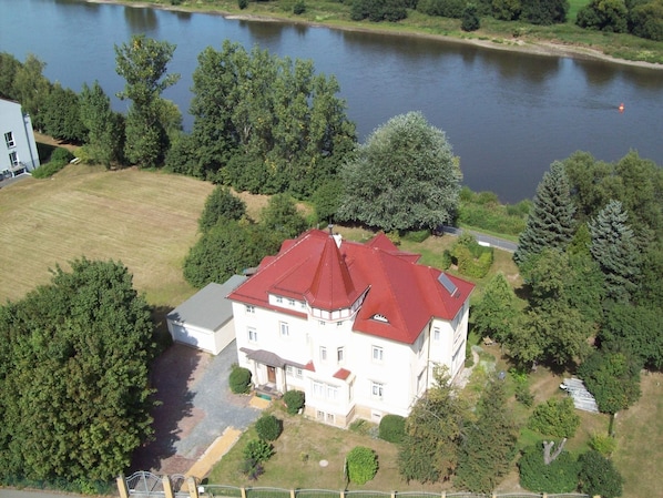 A large, multi-story building with a red roof, surrounded by a well-maintained lawn and trees, near a body of water. - Pension Villa Else (Heidenau)
