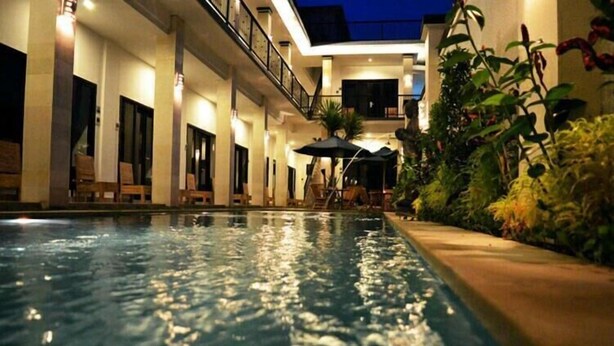 En swimmingpool med liggestole og parasoller, omgivet af beplantning og en bygning med balkon.