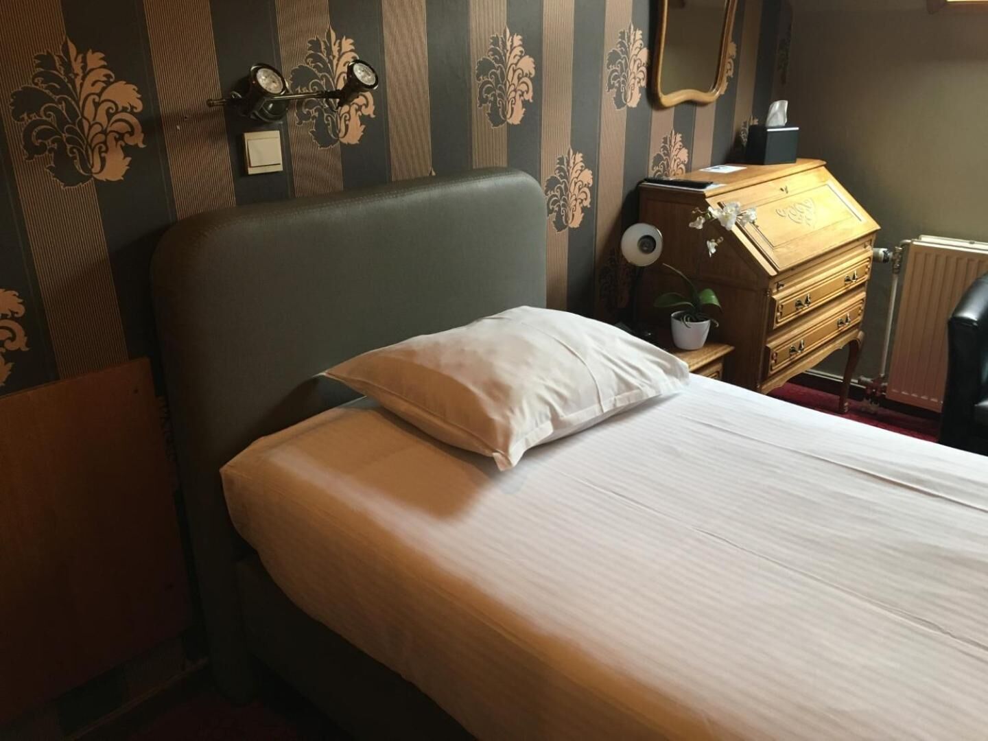 A neatly made bed with a pillow, a wooden dresser with a mirror, and a radiator in a room with striped wallpaper.
