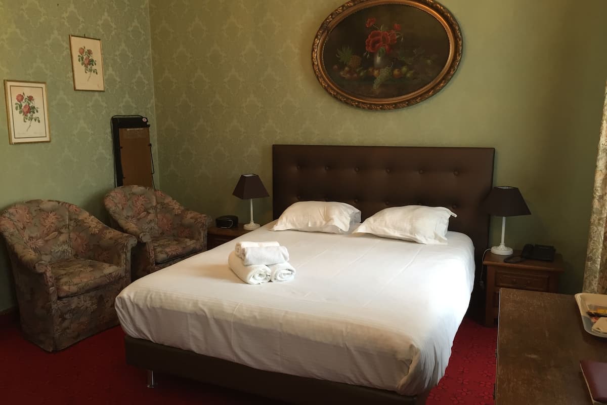 Hotellihuone, jossa on suuri sänky, kaksi nojatuolia, yöpöytä lampun kanssa, kehystetty kukkamaalaus ja punainen matto.