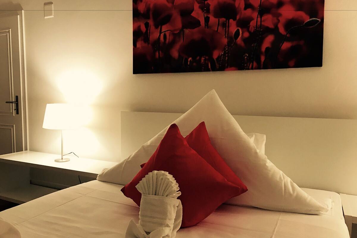 Una cama bien hecha con almohadas blancas y rojas, una toalla blanca y un cuadro colgado en la pared con flores rojas.